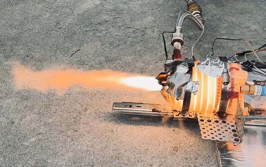 Orange flame streaming left from a resin engine wrapped in aluminum tape and clamped to a drawer rail on a gray concrete floor